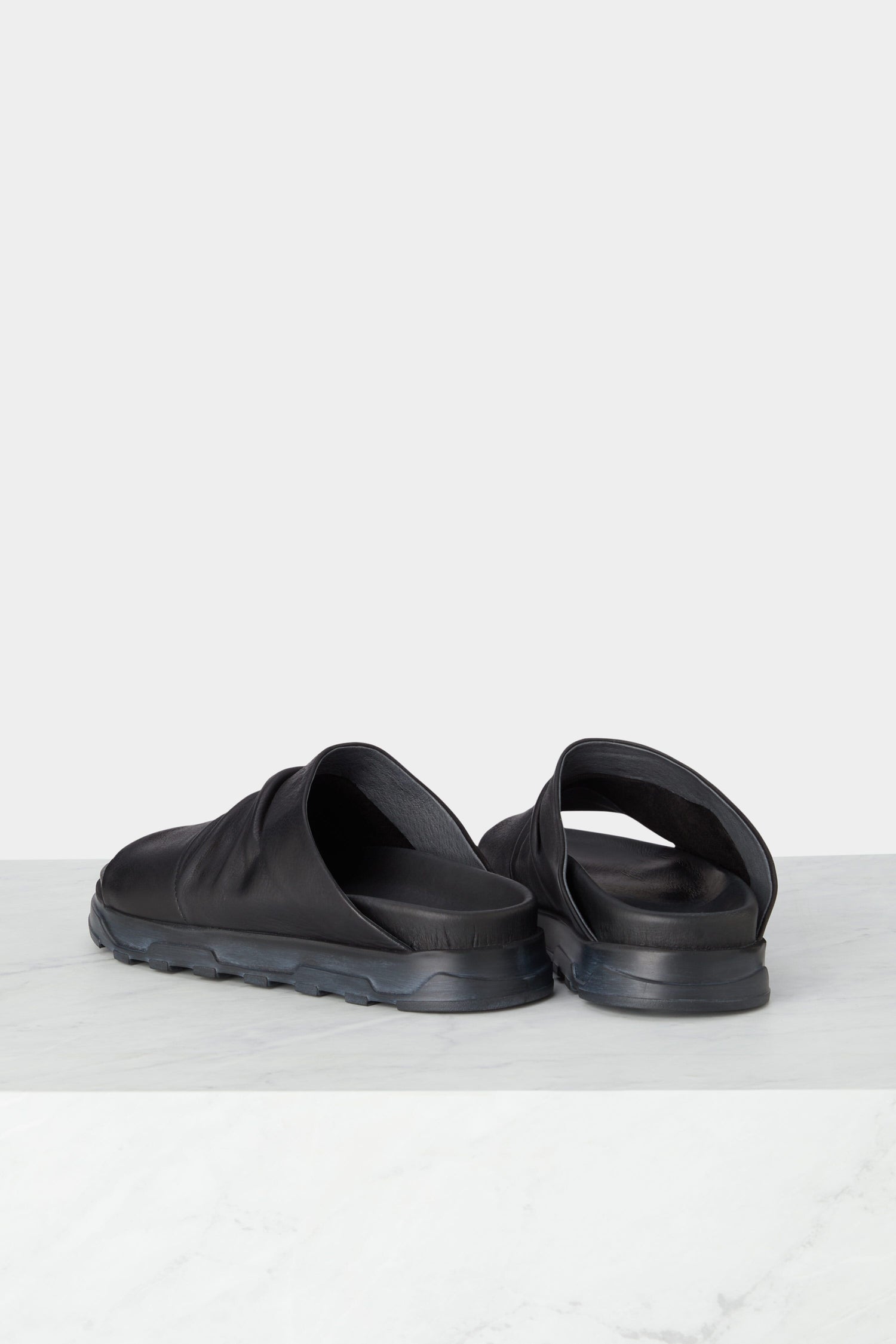 A pair of black Crushed Leather Sliders with textured soles, showcasing Scandinavian design, is displayed side by side on a light grey surface.