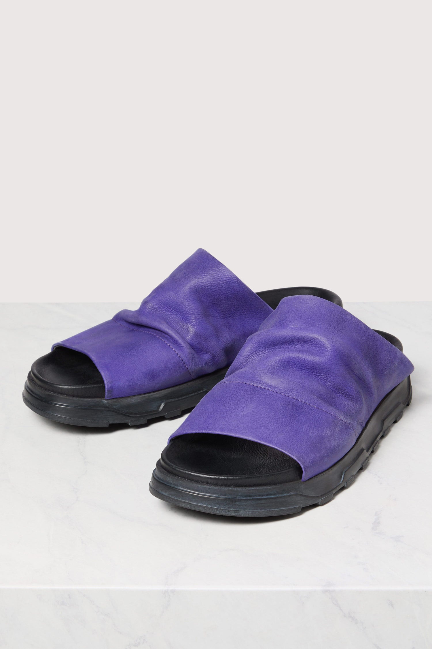 A pair of Crushed Leather Sliders in purple on a marble surface.