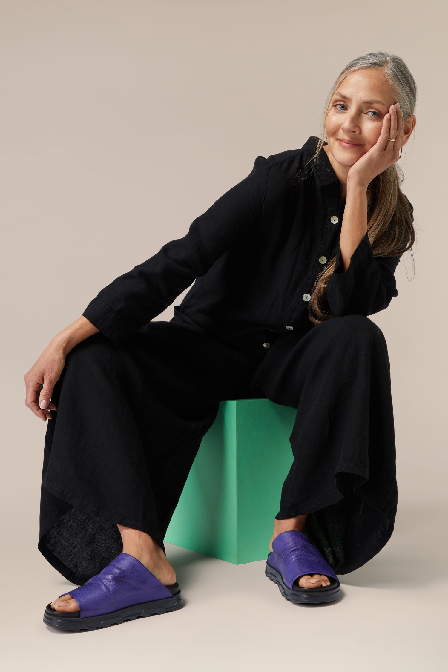 A woman in a black outfit sitting on a green stool against a neutral background, with one hand resting on her cheek and wearing Crushed Leather Sliders.