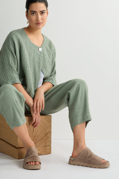 A person sits on a wooden block, dressed in a light green outfit and chic Biscotti Crushed Leather Sliders against a plain background.