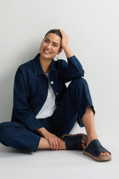 In a navy outfit and Crushed Leather Sliders, this person radiates effortless luxury. Smiling with one hand on their head, the cushioned soles of their sandals provide comfort against the plain backdrop.