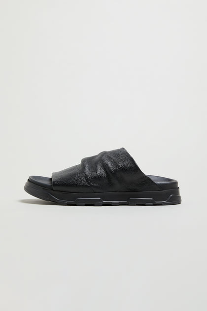 A single Crushed Metallic Leather Slider in black, featuring a textured leather upper and thick, ridged rubber sole, is shown in side profile against a plain light background.