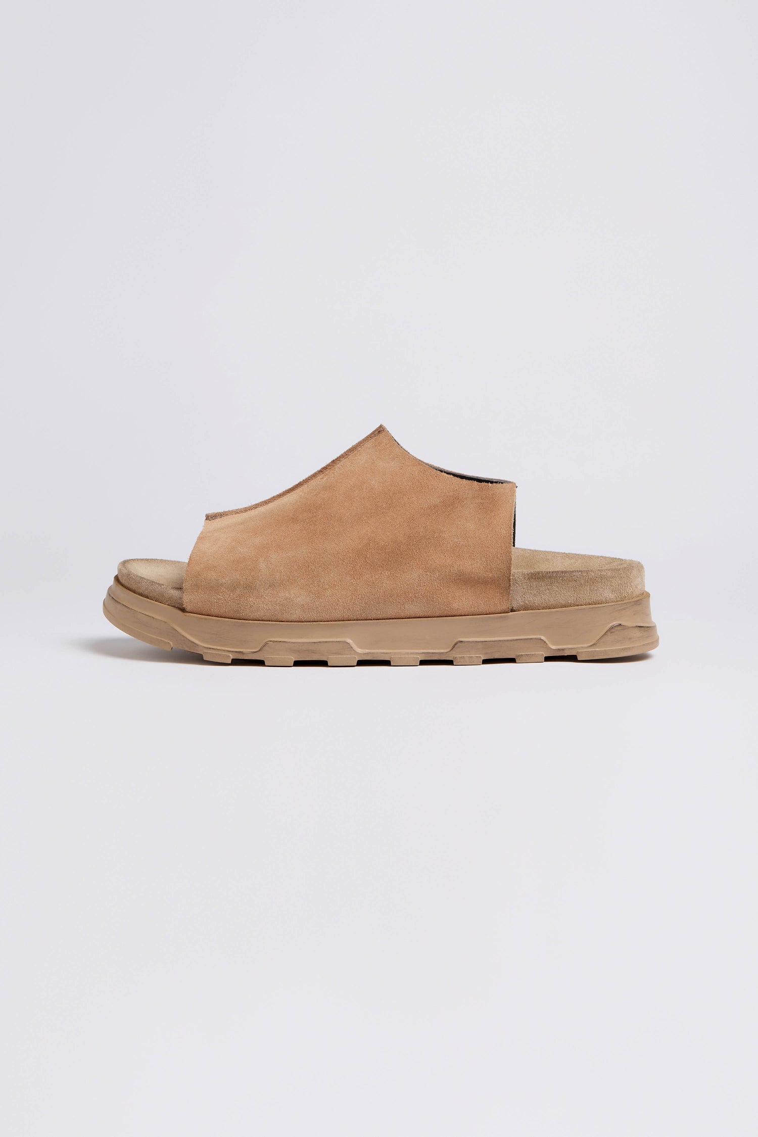 A side view of the Seam Suede Sliders, a brown suede slip-on sandal showcasing contemporary minimalism. It features a textured beige rubber sole and is handcrafted in Italy, set against a plain white background.