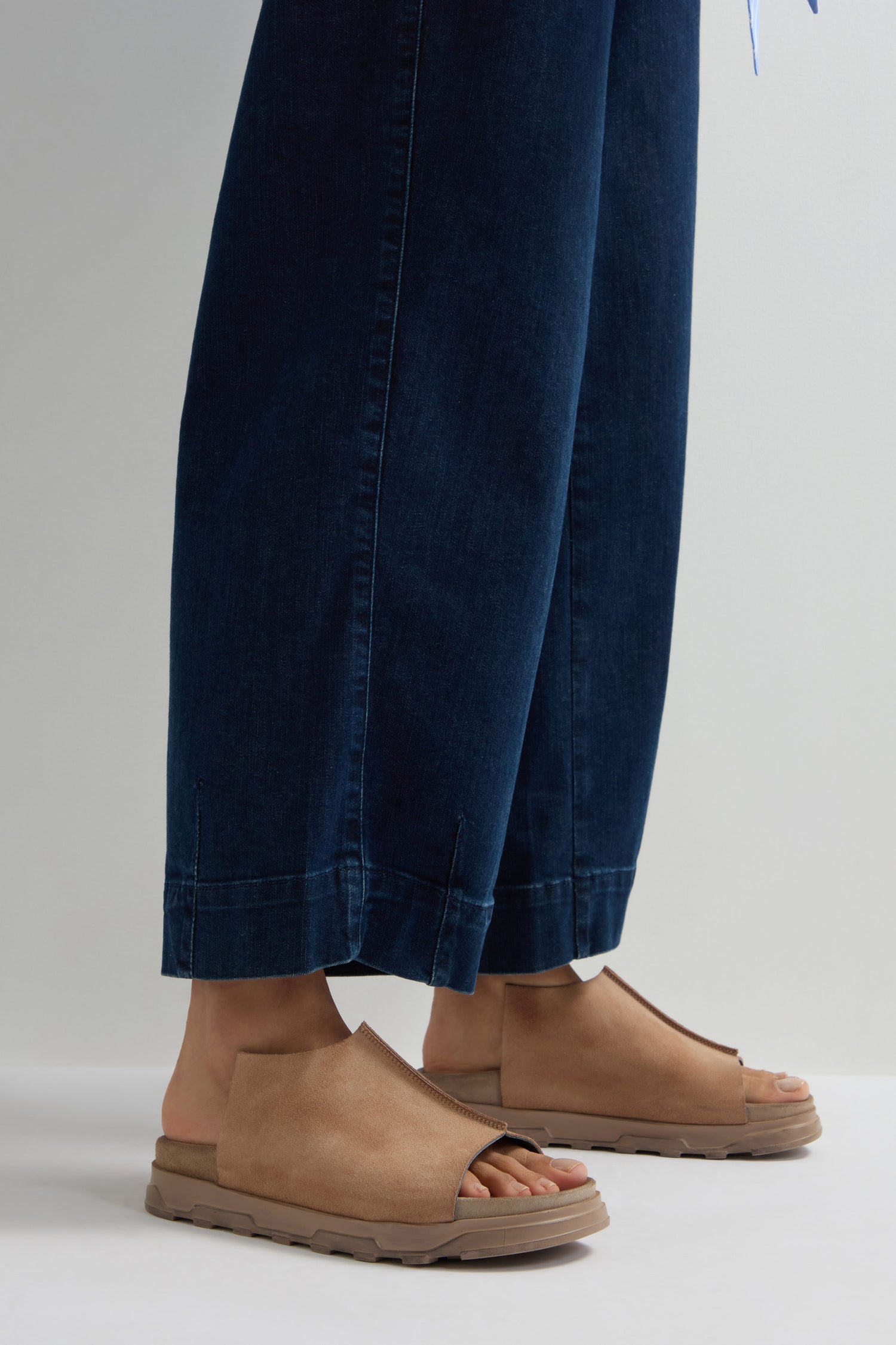 A person stands on a light surface in dark blue wide-leg pants with Seam Suede Sliders, tan open-toe sandals handcrafted in Italy, embodying contemporary minimalism.