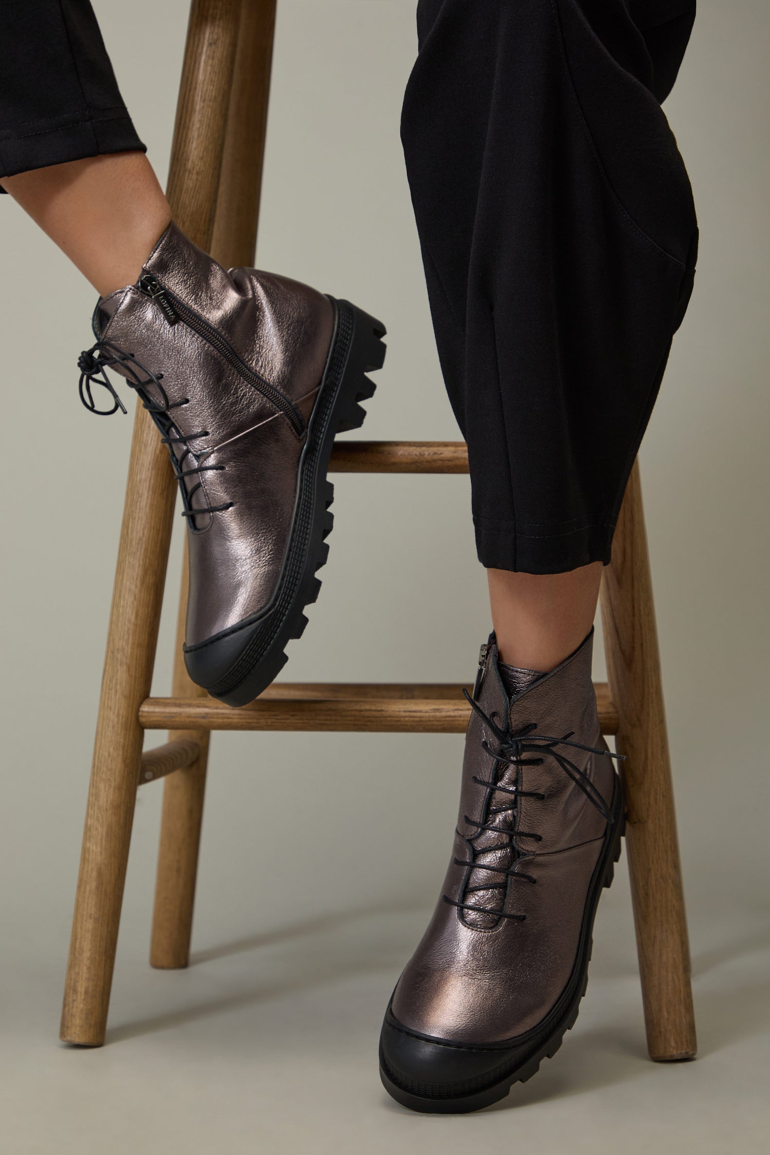 A person wearing Rose Metallic Lace Up Leather Boots with black soles and cropped black pants is seated on a wooden stool.