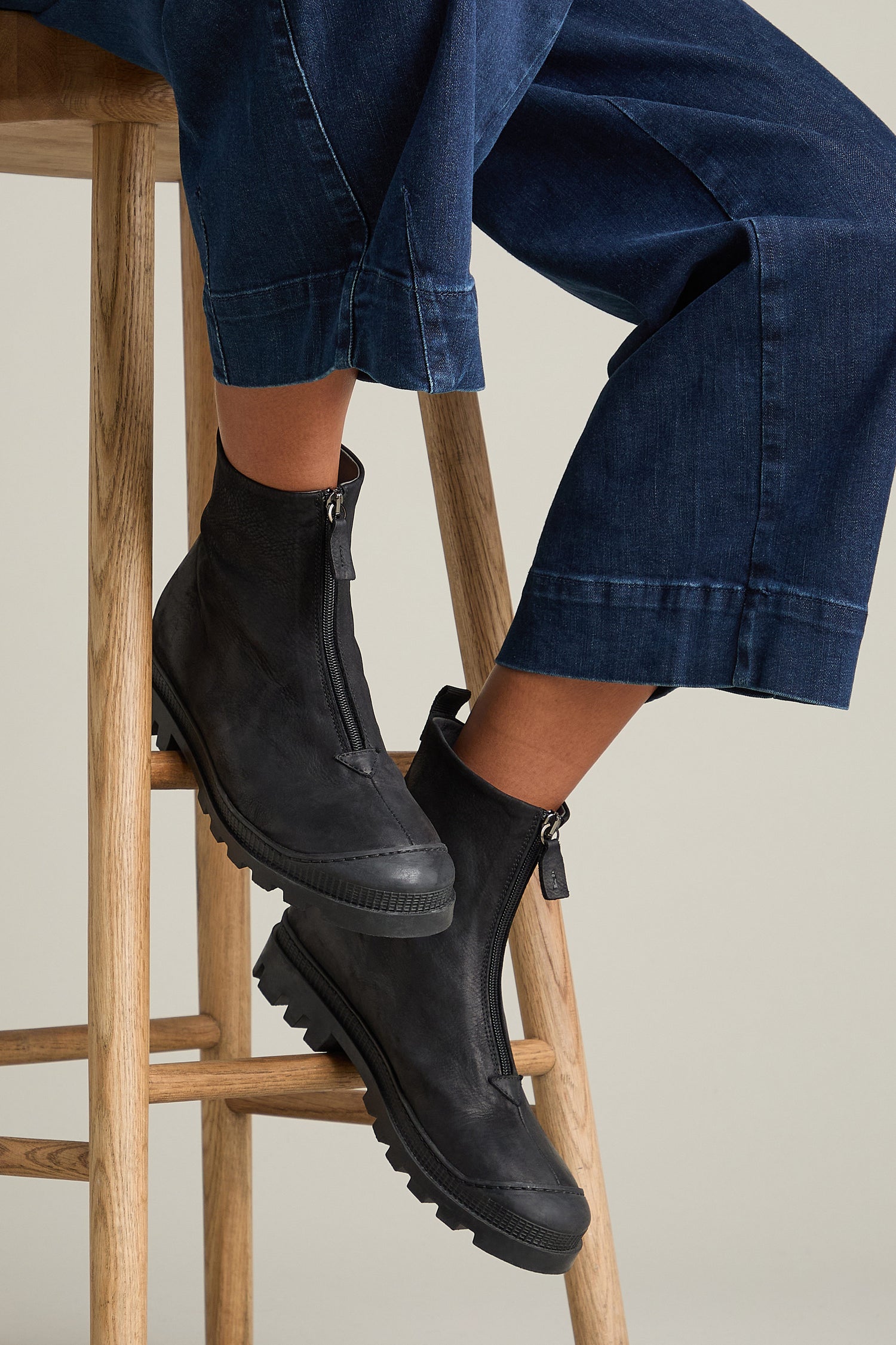 Person sitting on a wooden stool, wearing dark denim pants and sleek black Zip Front Leather Boots with sturdy calfskin soles.