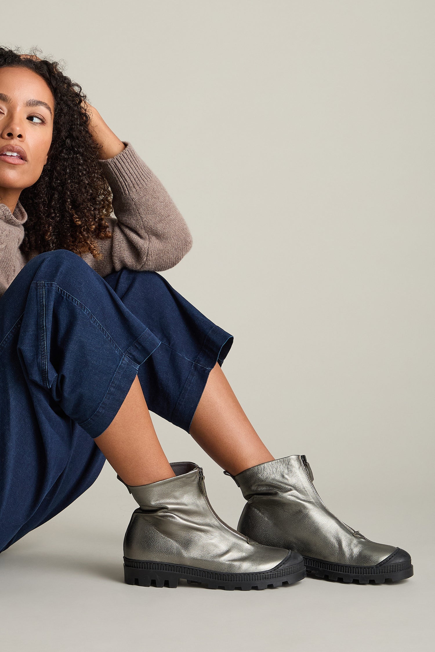 A person in a brown sweater and blue pants sits on the floor, wearing Zip Front Leather Boots in metallic gray crafted from calfskin with black soles.