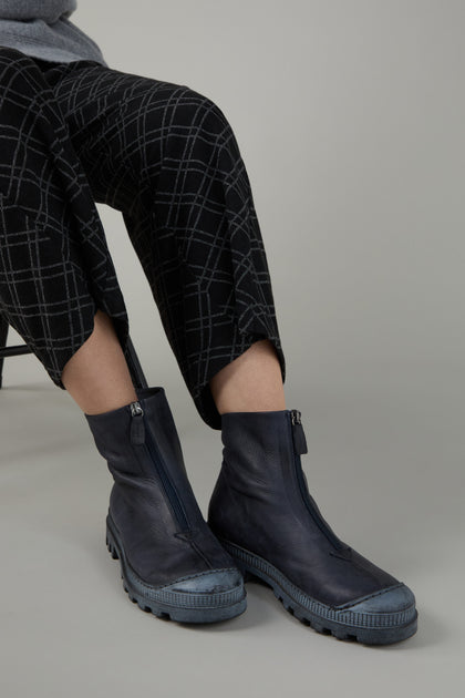 A person in black patterned pants wears Navy Zip Front Leather Boots while seated on a chair against a neutral gray background.