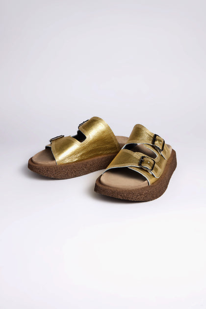 These exclusive Metallic Gold Double Buckle Leather Sliders from Sahara are handcrafted in Italy, featuring two buckled straps and cork soles on a white backdrop.