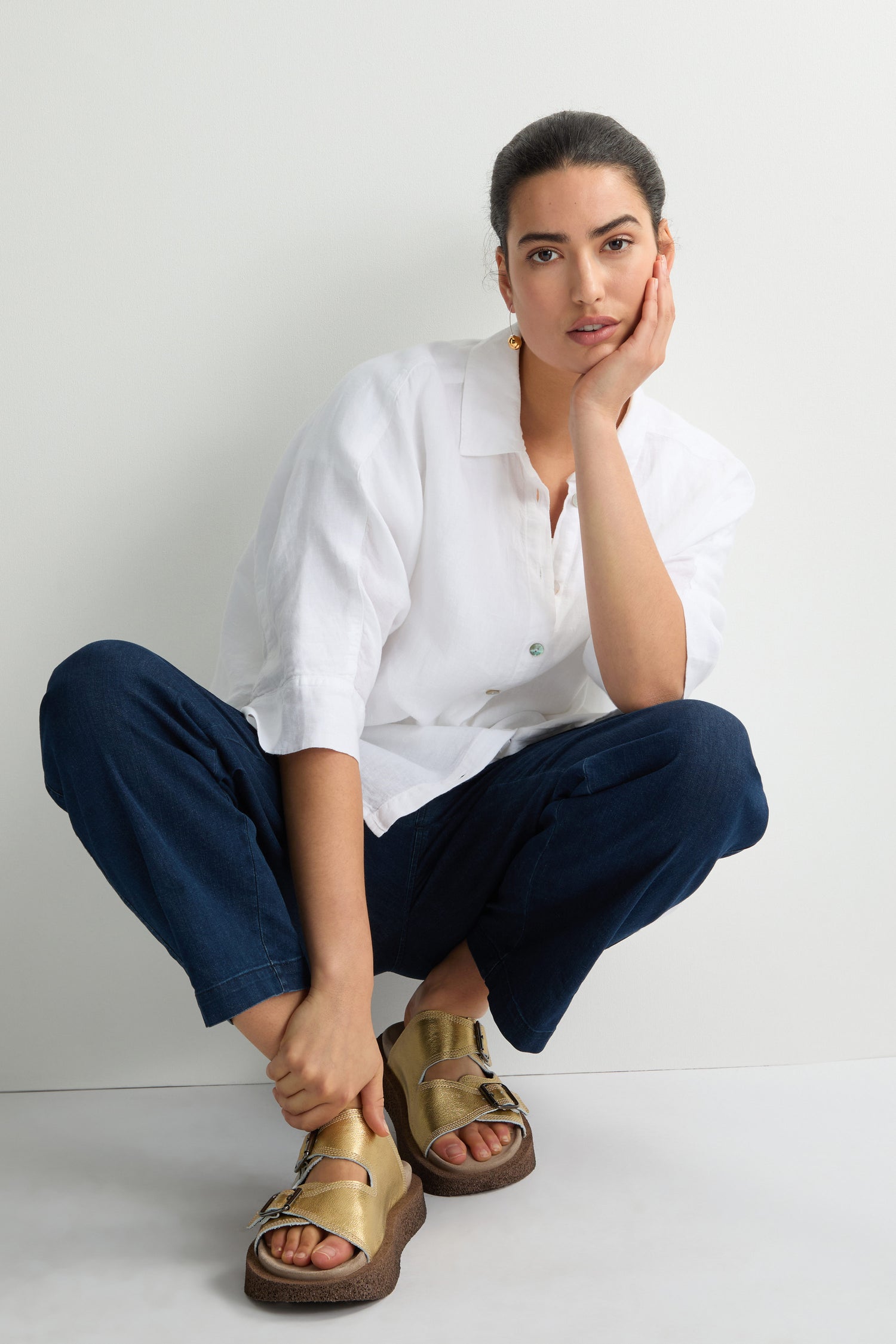 A person crouches against a white wall, wearing a white shirt and dark blue pants. Their feet feature Metallic Gold Double Buckle Leather Sliders, handcrafted in Italy.