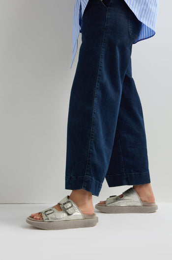 Person wearing wide-leg jeans, a striped shirt, and Sahara exclusive Metallic Silver Double Buckle Leather Sliders standing against a white background.