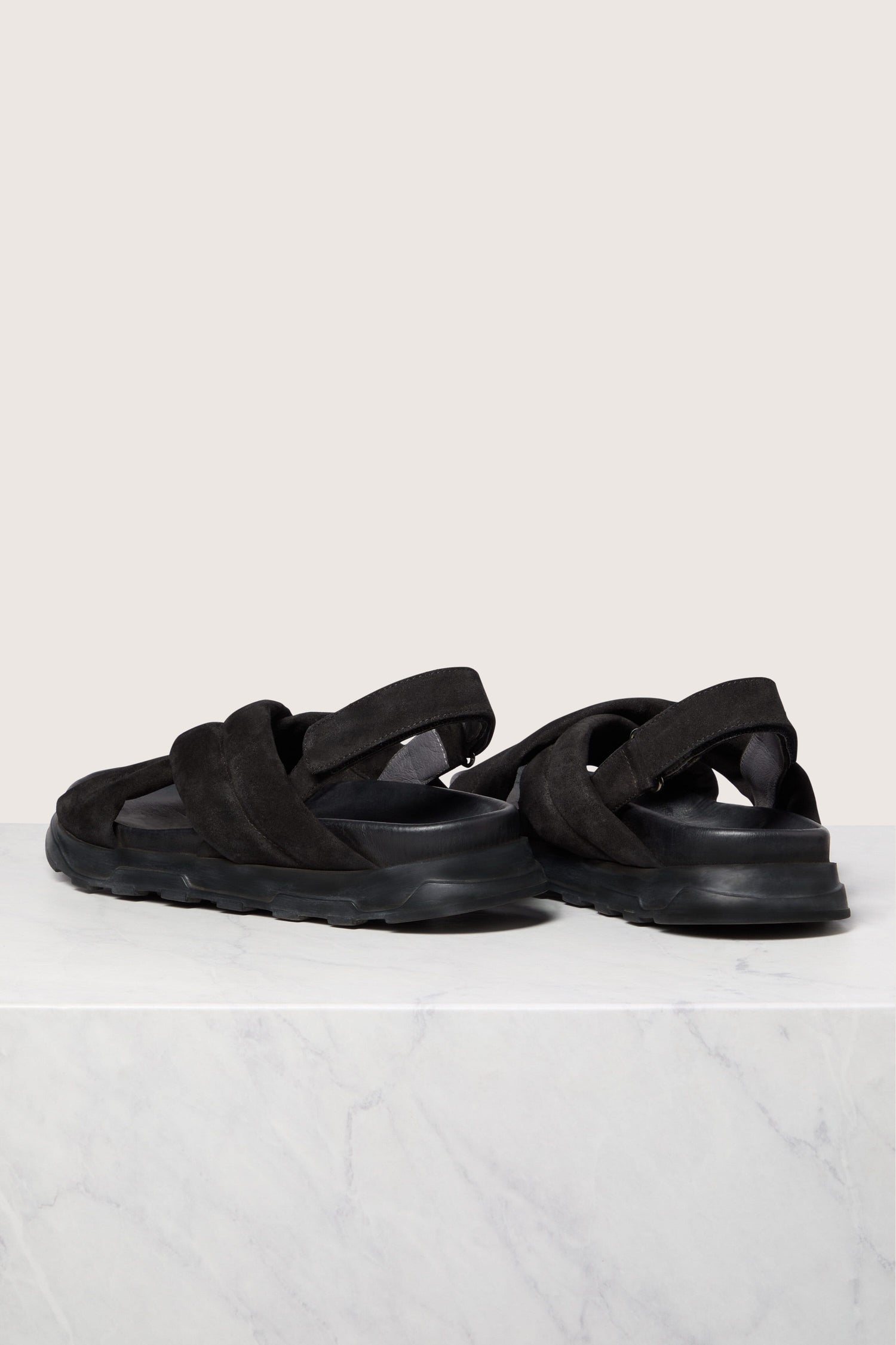 A pair of black 'Suede Crossover Sandal' with velcro straps and a handmade sole on a white marble surface.