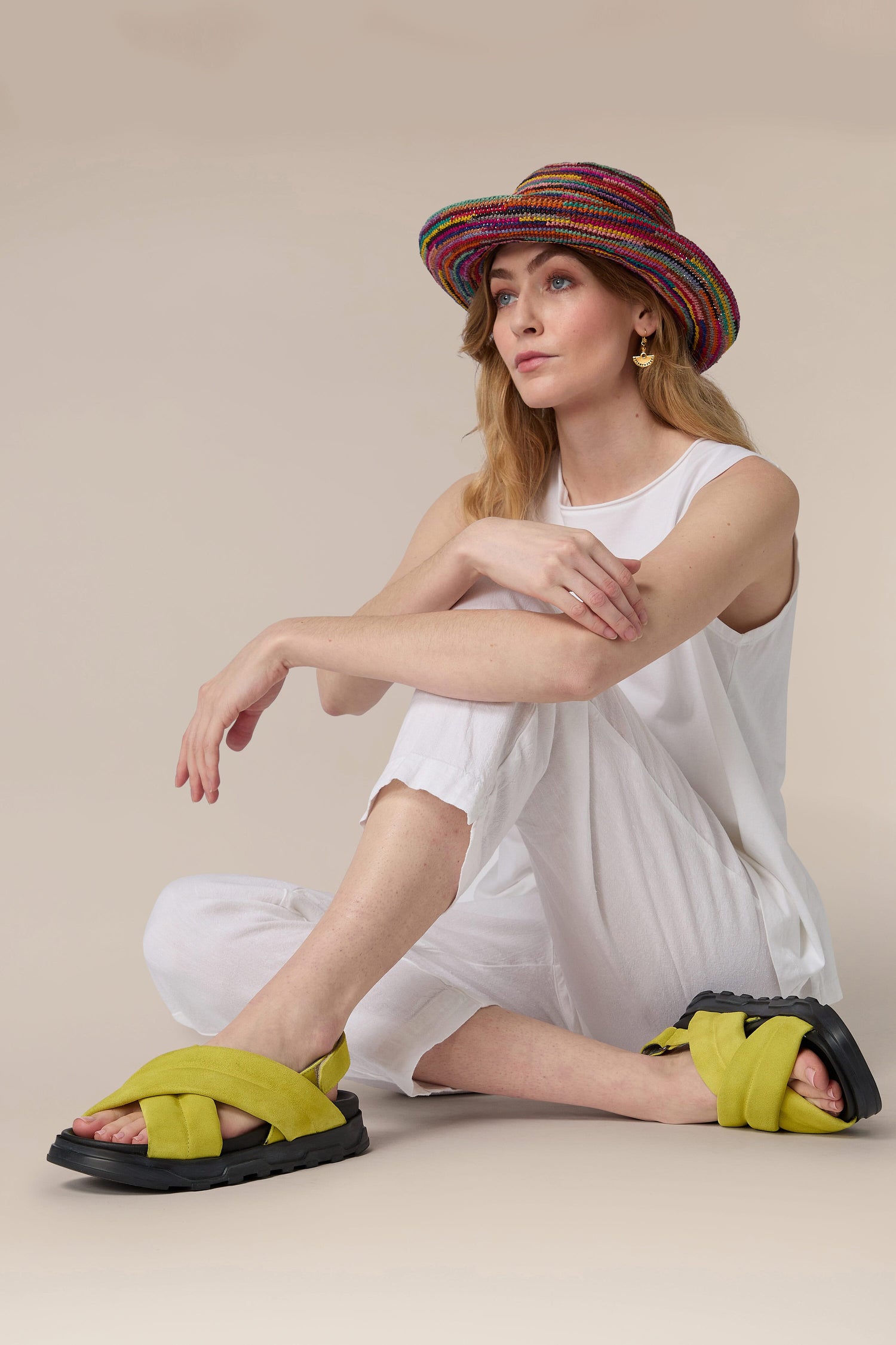 Woman sitting on the floor wearing a white outfit, colorful hat, and yellow Suede Crossover Sandal.