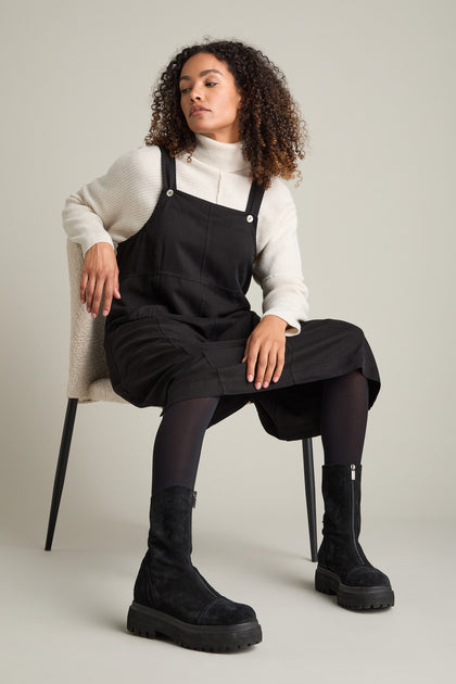 A person with curly hair sits on a chair wearing a white turtleneck, black overalls, black tights, and stylish Zip Front Suede Platform Boots. They look to the side with a relaxed posture.