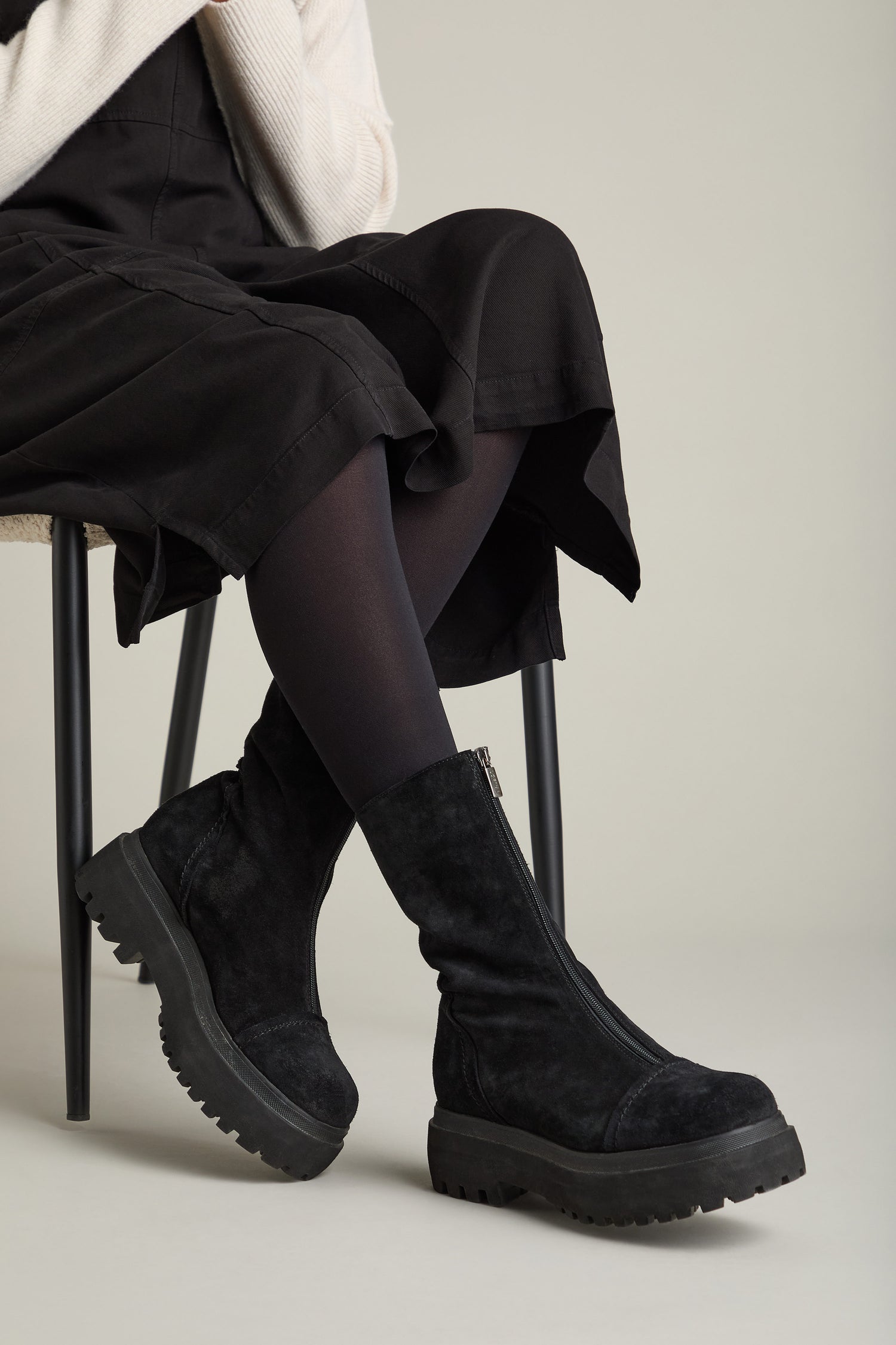 Person sitting on a chair wearing a black skirt, black stockings, and black Zip Front Suede Platform Boots.