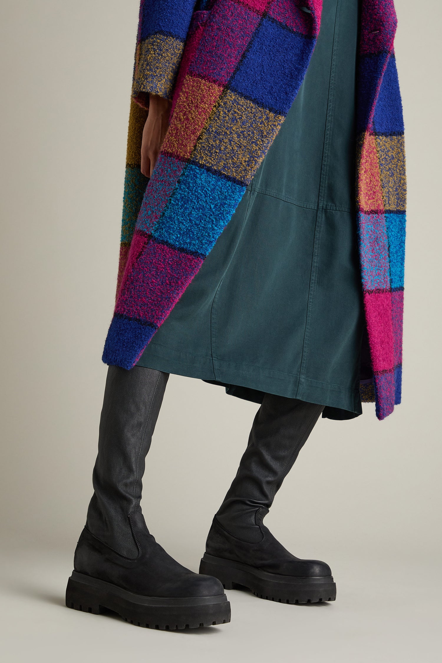 A person wearing a colorful checkered coat, a teal dress, and black Long Stretch Knee High Leather Boots made of calfskin leather stands against a plain background.