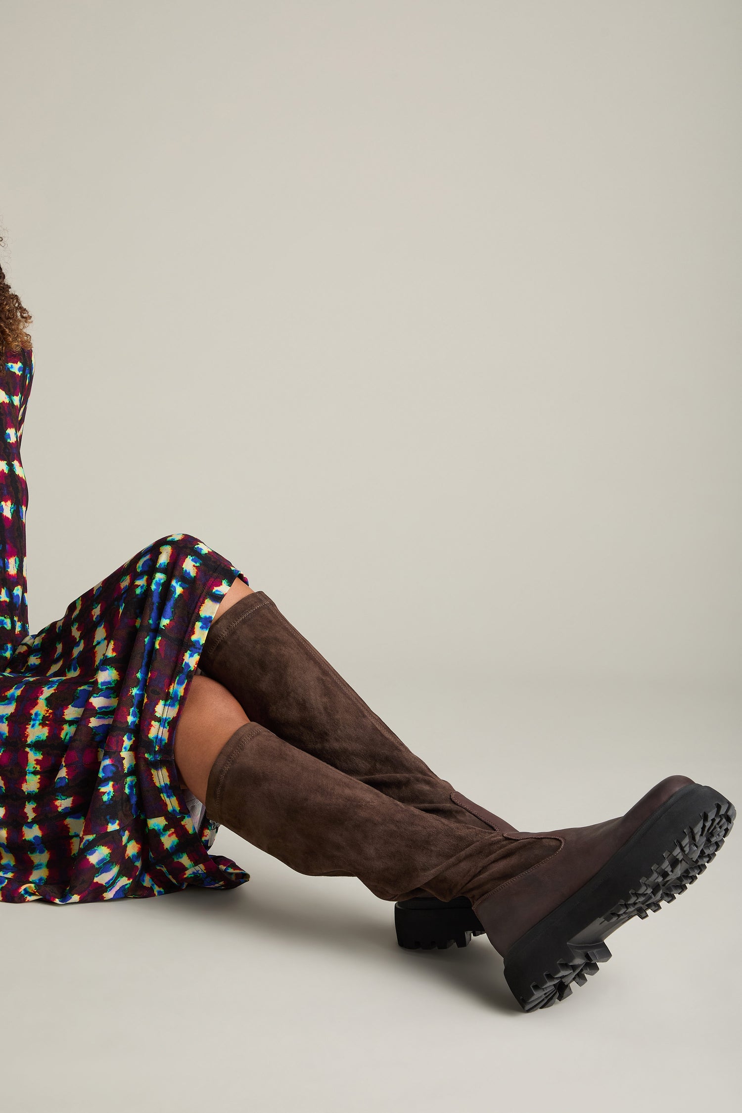 Person wearing Long Stretch Knee High Leather Boots made of calfskin leather and a colorful patterned dress, seated against a plain background.