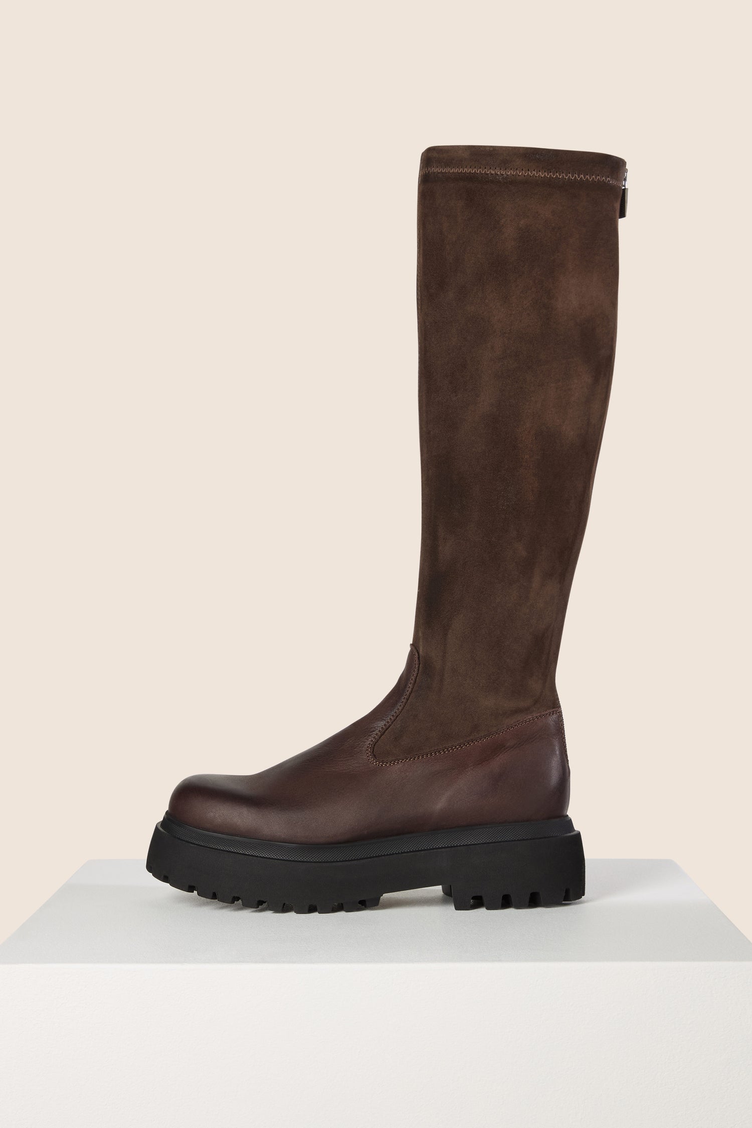 The Long Stretch Knee High Leather Boots are a stylish and chic tall boot crafted from brown calfskin leather and suede, featuring a chunky black sole. These boots are elegantly showcased on a white platform against a beige background.