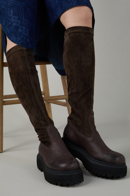 A person sits on a wooden chair, legs slightly apart, wearing Chocolate Long Stretch High Knee Suede Boots with a thick platform sole and a blue skirt visible.