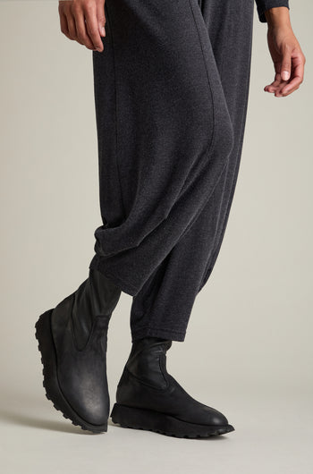 A person is showcasing their lower legs, dressed in loose-fitting dark gray pants and a pair of Leather Stretch Boots made from black calfskin with hand-cut soles, all set against a minimalist backdrop.