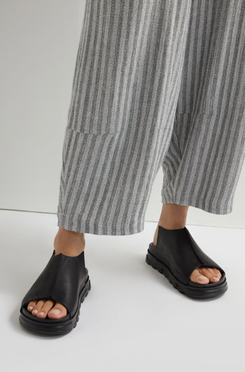 Wearing Seam Leather Sliders in sleek black, complemented by gray striped pants, a person effortlessly stands on a light backdrop, embodying modern minimalism.