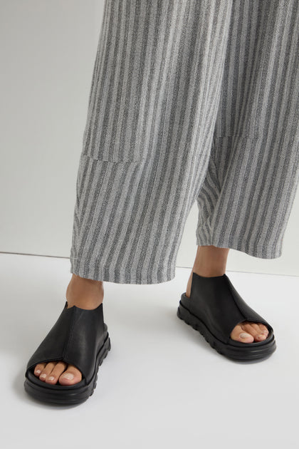 Wearing Seam Leather Sliders in sleek black, complemented by gray striped pants, a person effortlessly stands on a light backdrop, embodying modern minimalism.