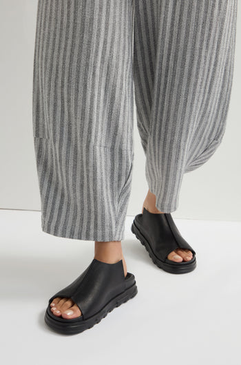 A person exudes modern minimalism with gray striped pants and sleek black Seam Leather Sliders, showcasing their chic open-toe design.