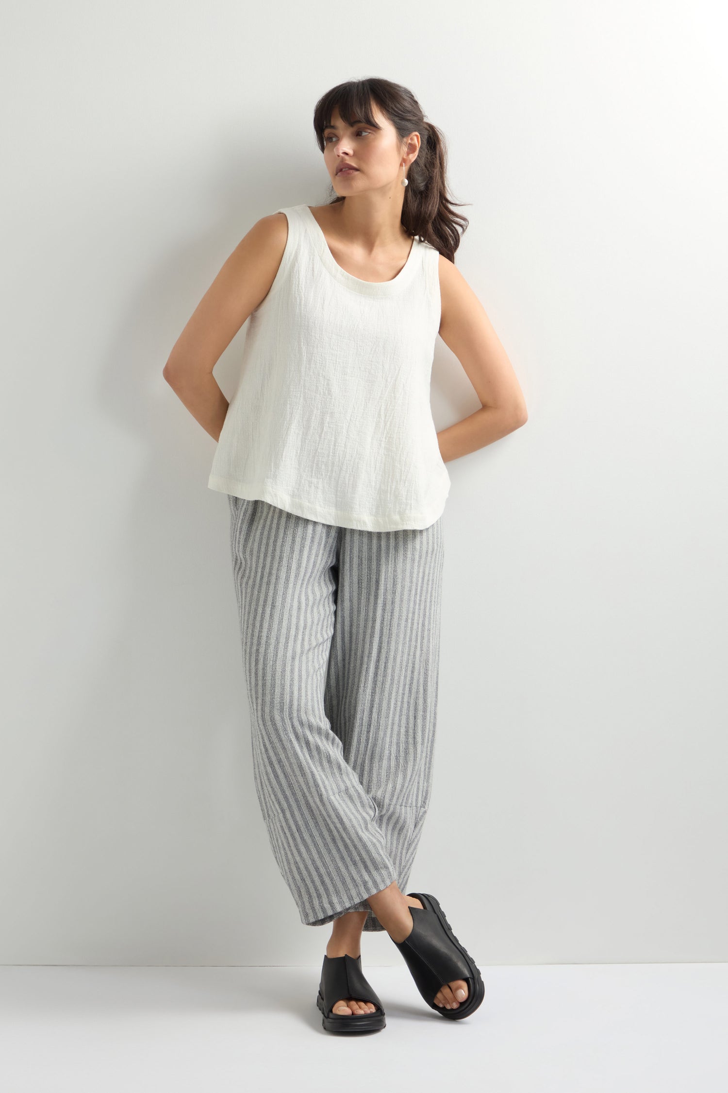 A person embodies modern minimalism, donning a white sleeveless top with gray striped pants and sleek Seam Leather Sliders. They stand against a light-colored wall, perfectly capturing understated sophistication with their chic ensemble.