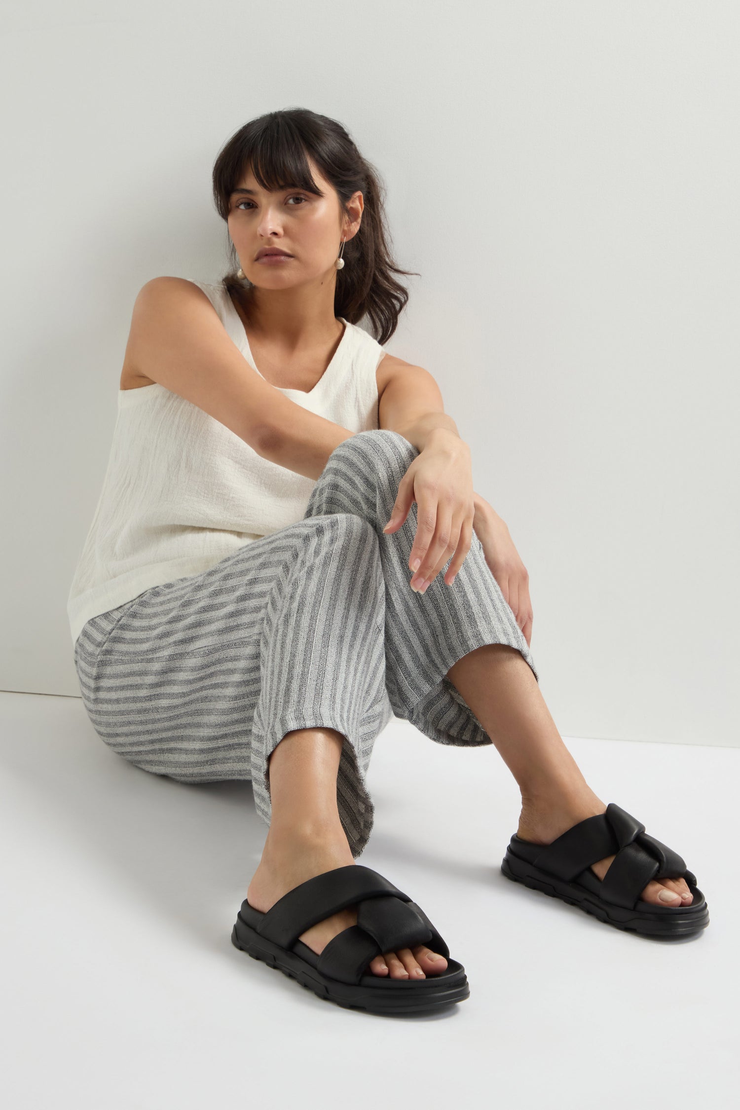 A person sits on the floor against a white wall, dressed in a sleeveless white top, gray striped pants, and sleek Lattice Leather Sliders.