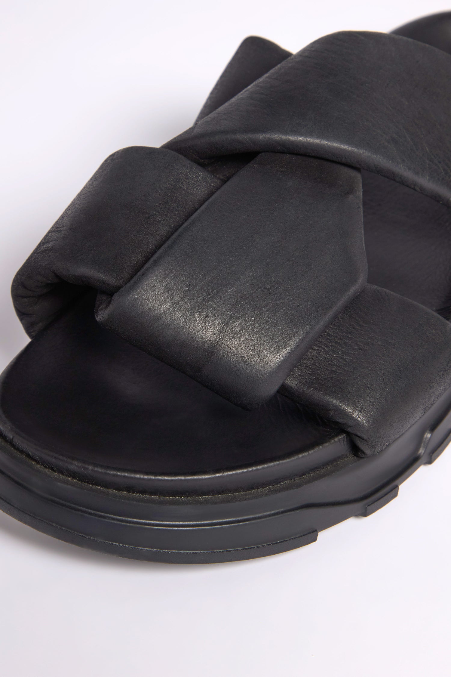 Close-up of Lattice Leather Sliders in black highlights wide calfskin cross-over straps and a thick sole on a plain white background, embodying sleek contemporary style.