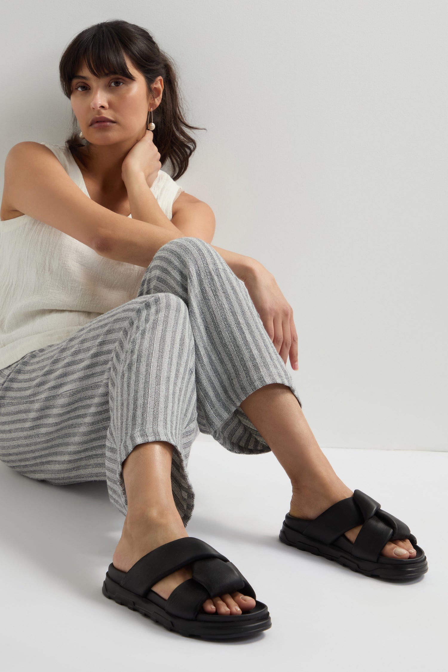 A person sits on the floor against a white wall, wearing a sleeveless white top, striped pants, and Lattice Leather Sliders.