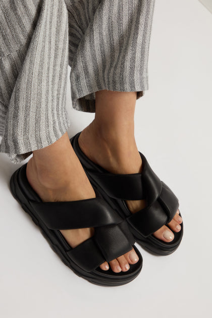 A person is wearing striped pants and chic black Lattice Leather Sliders, reminiscent of the elegance found in Lofina shoes.