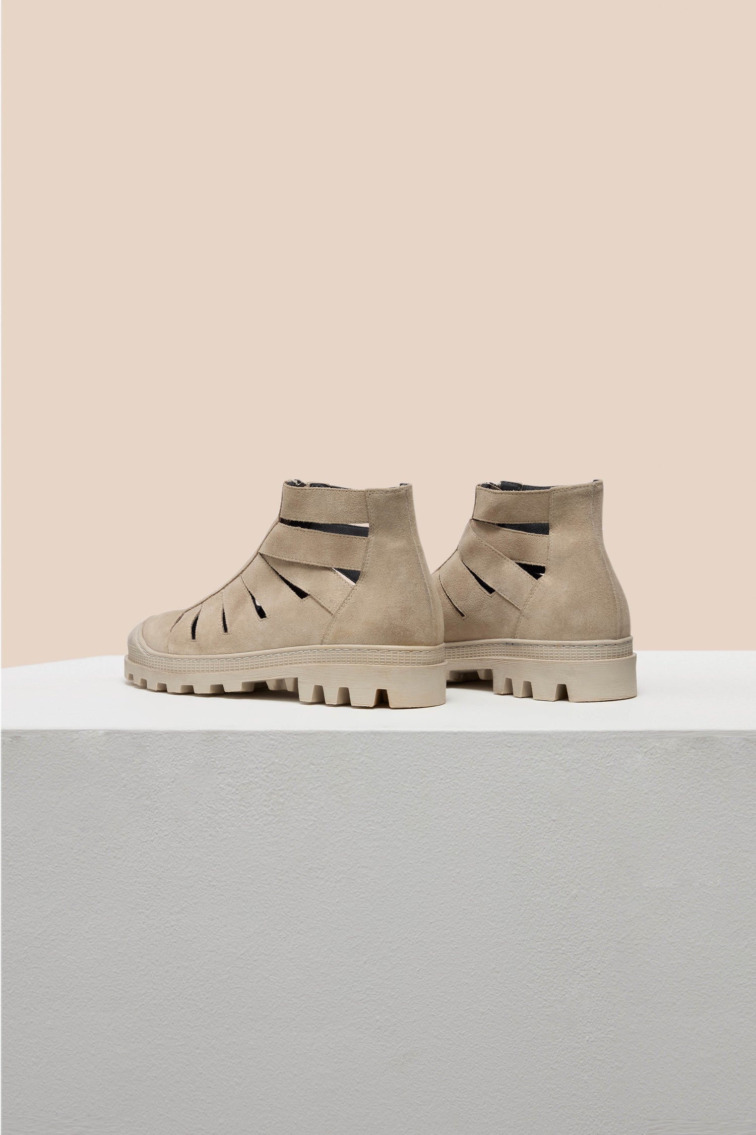 Beige Cut Out Suede Zip Boots with black accents, featuring an edgy design and rugged rubber sole, rest on a white platform against a light peach background.