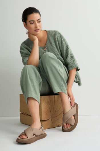 A woman in a textured green outfit and Crossover Leather Sandals featuring a cushioned footbed sits on a wooden block against a light background, showcasing Scandinavian craftsmanship.