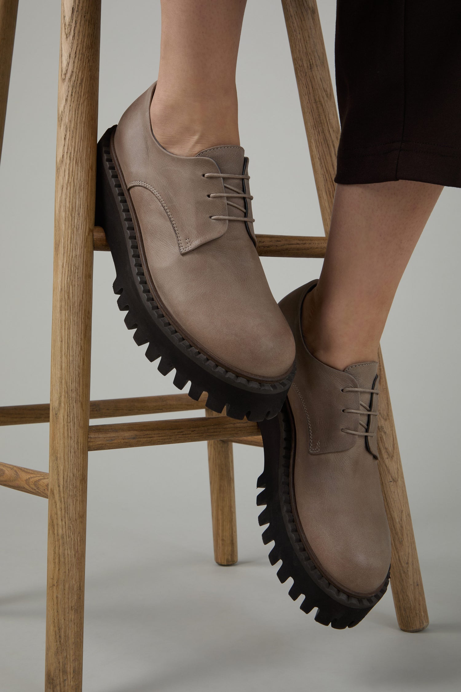 Wearing Two Tone Lace Up Leather Brogues with thick black ridged soles, the person rests their feet on a wooden stool's footrest.