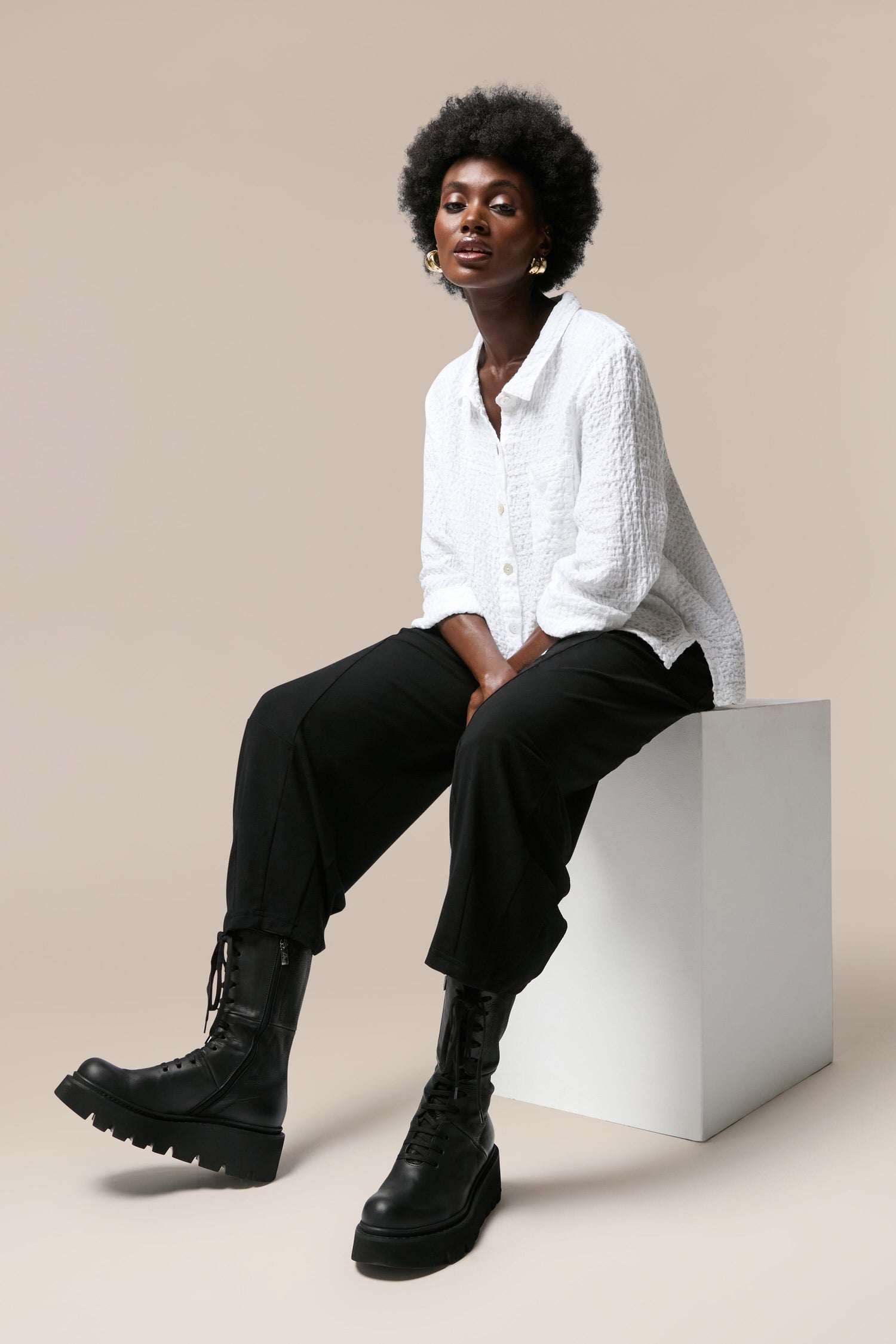 A black woman in a white shirt and black pants sitting on a white cube, showcasing Platform Lace Up Leather Boots.