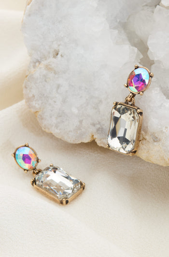 The Crystal Gemstone Earrings feature iridescent oval stones and rectangular gemstones set in a gold-tone frame, elegantly displayed on a white crystalline rock and beige fabric.