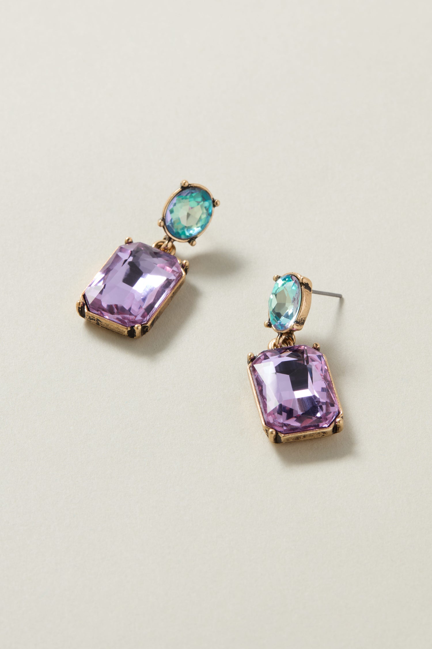 The Twin Gemstone Earrings, featuring lilac and azure rectangular gemstones with round blue gemstones set above them, are displayed on a light-colored surface.