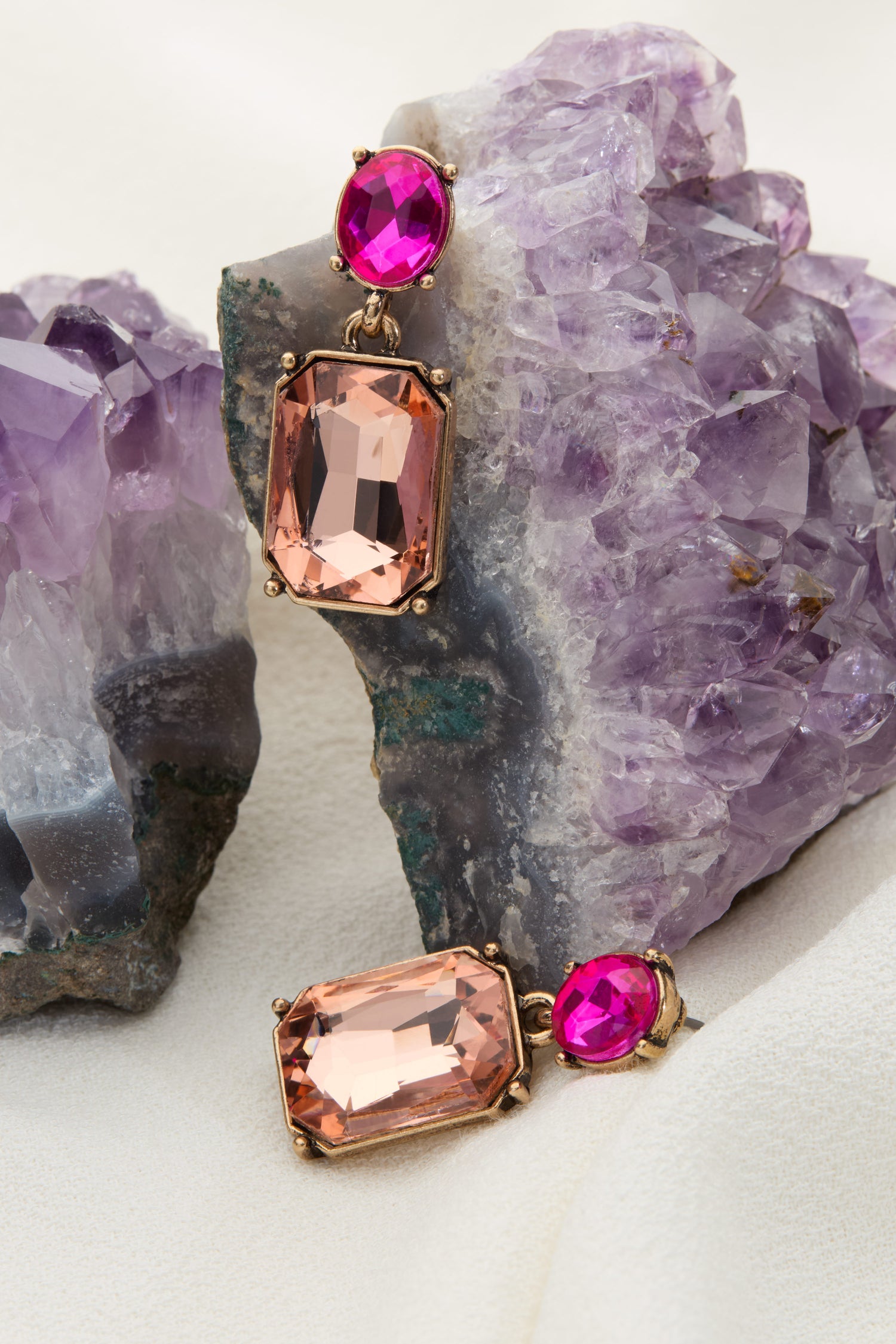 The Twin Gemstone Earrings, featuring pink and magenta gemstones, are displayed against a backdrop of lilac and azure amethyst crystals.