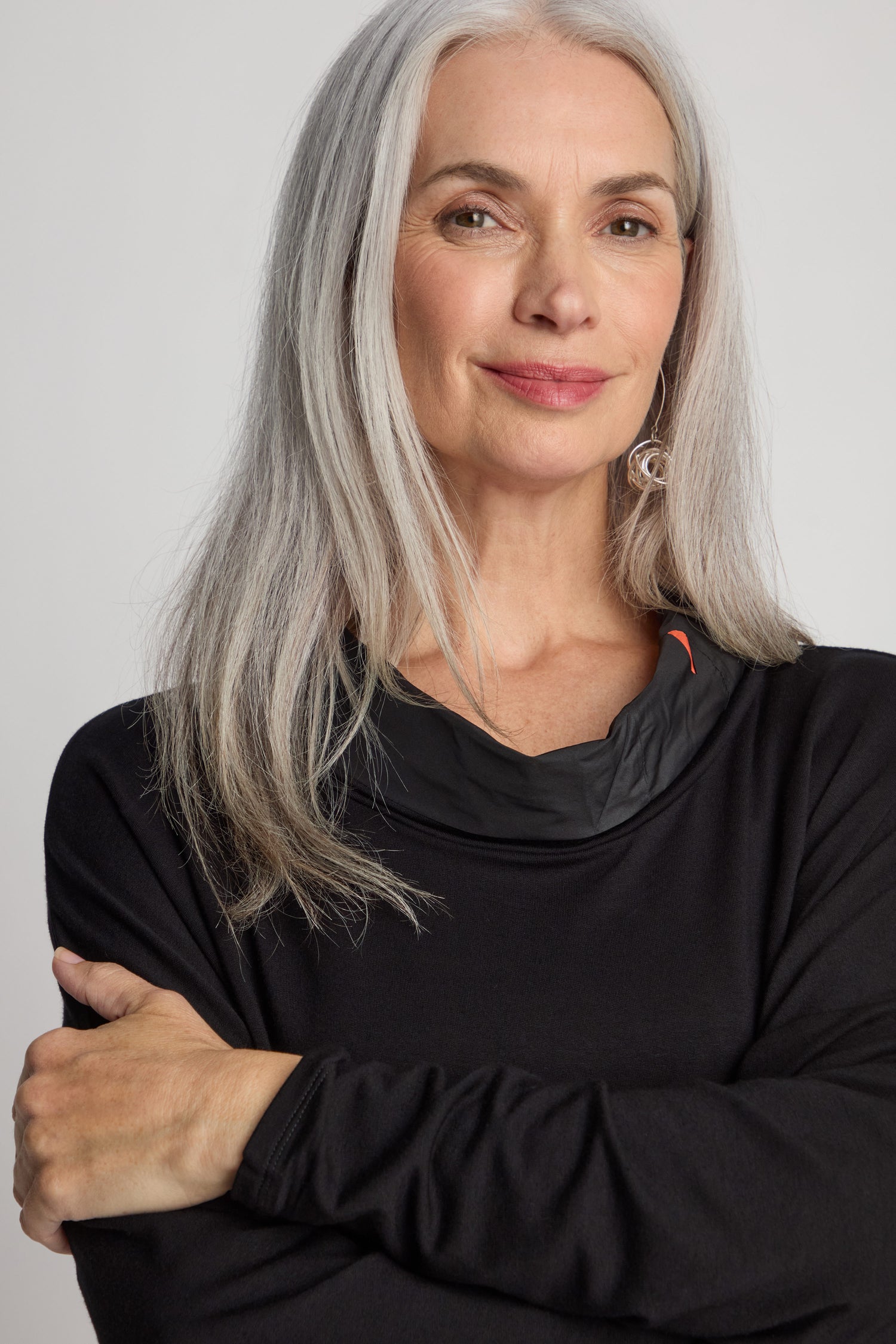 A woman with long gray hair wearing a black Cut Out Cowl Neck Tunic stands with her arms crossed, smiling slightly.