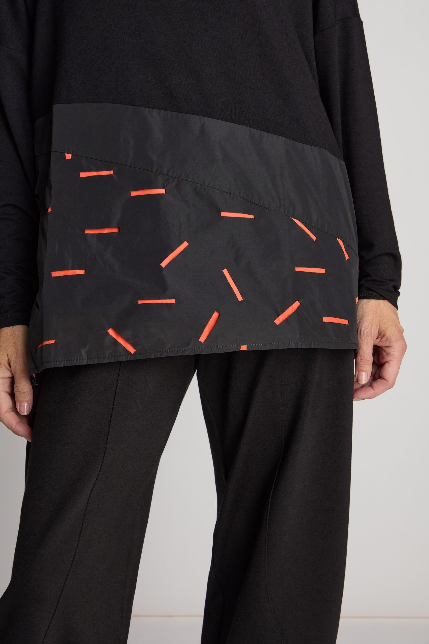 The person is wearing a Cut Out Cowl Neck Tunic paired with black pants, featuring a minimalist design and contrasting fabric sections near the hem in striking black and red patterns.