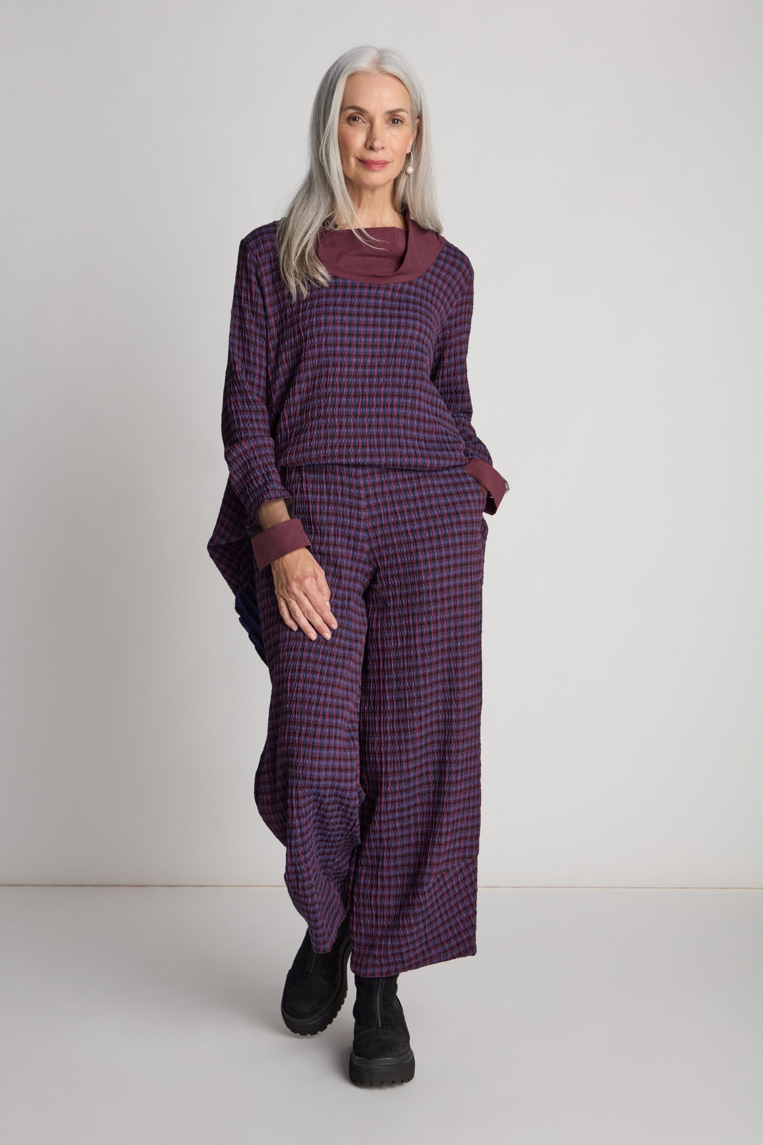 A woman with long gray hair showcases the Check Trouser, a stylish pair of wide-leg trousers in a purple and red checkered pattern, paired with a matching long-sleeve top and black shoes. She stands against a light gray background, embodying the essence of sustainable fashion.