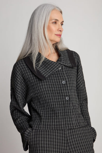 A person with long gray hair is wearing the Check Shawl Collar Jacket, a textured dark gray buttoned garment featuring a check shawl collar, and is standing against a plain background.