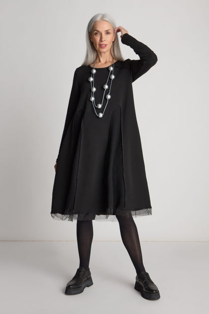 An older woman with long gray hair stands in a black Godet Net Hem Jersey Dress by Luukaa, adorned with a necklace featuring large beads. She completes her outfit with black tights and black shoes.