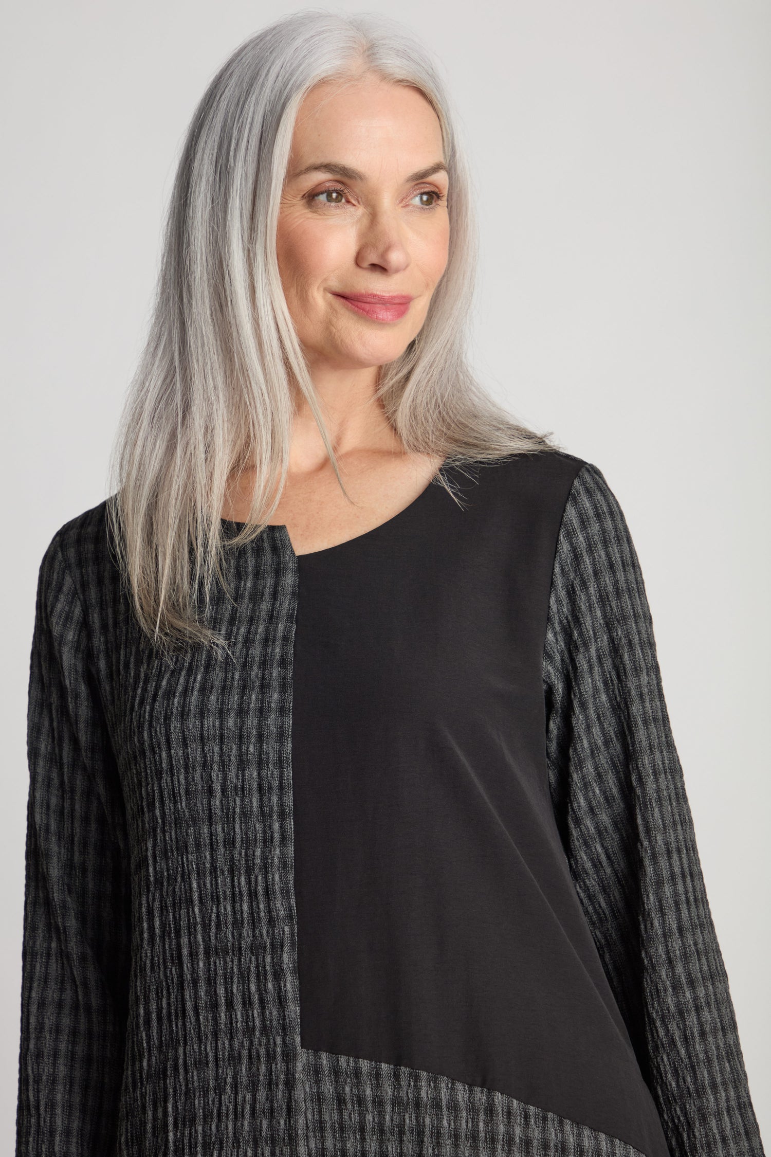 A woman with long gray hair stands against a plain background, wearing the Patched Check Dress, a black and gray textured long-sleeve top that features a subtle contrasting patchwork design.