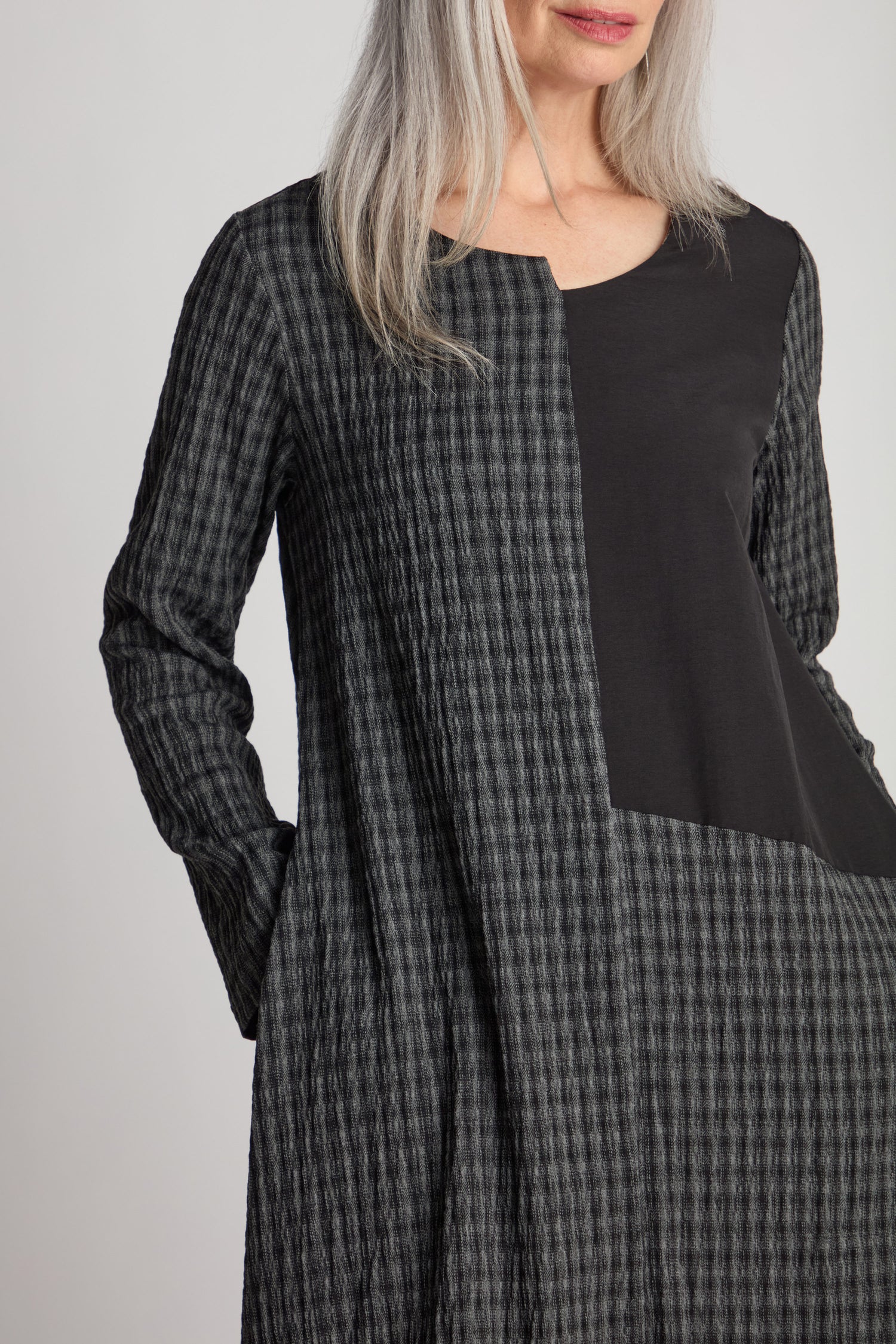 A person with long gray hair is wearing a Patched Check Dress, which features a long-sleeved, black and gray checkered design with an asymmetric hem. The photo is cropped to show the upper torso and part of the head.