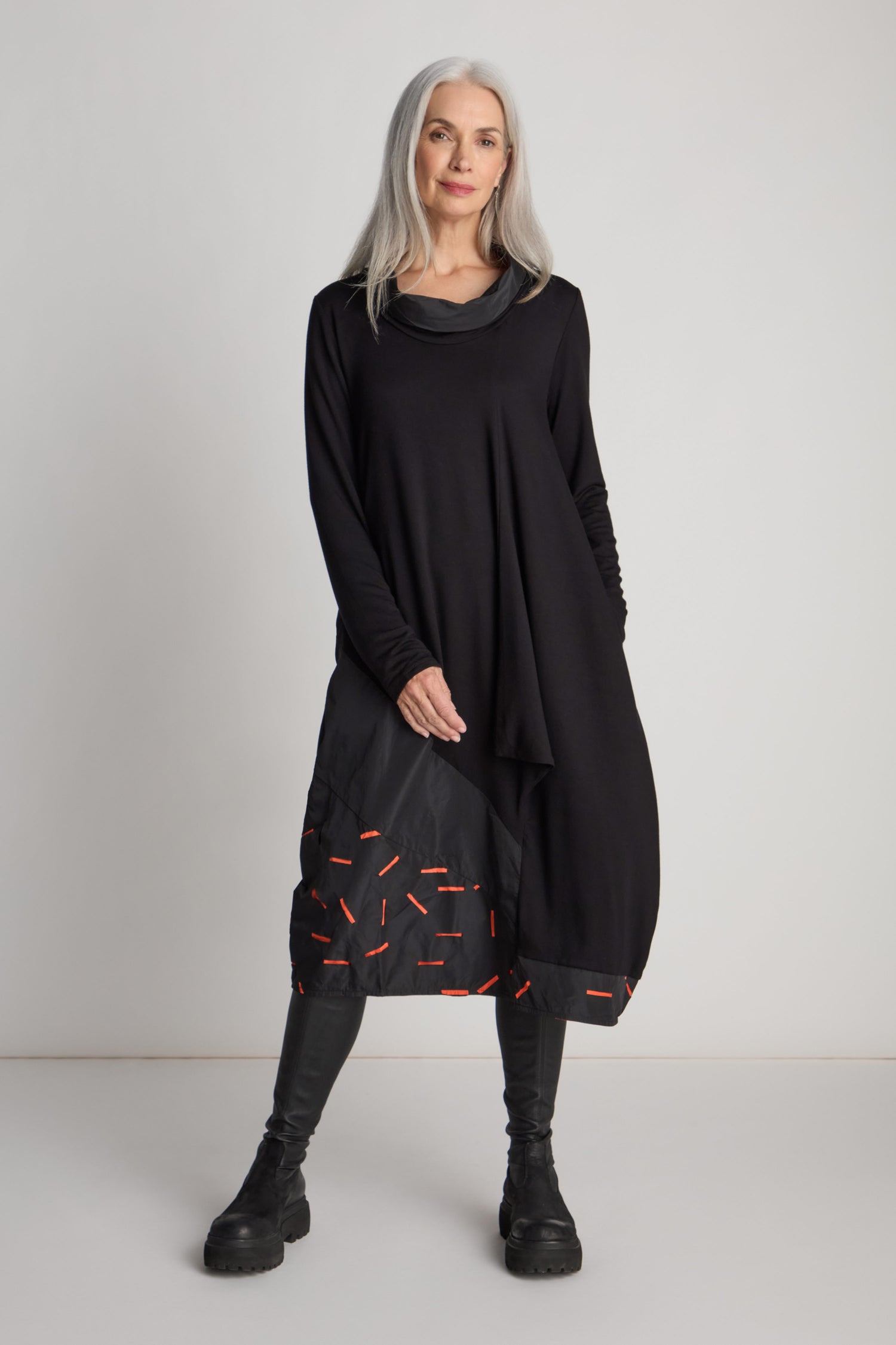 A woman with long grey hair is standing in a stunning Cut Out Cowl Neck Dress by Luukaa, a true statement piece with red accents near the hem. She pairs it effortlessly with black leggings and boots, all set against a plain white background.