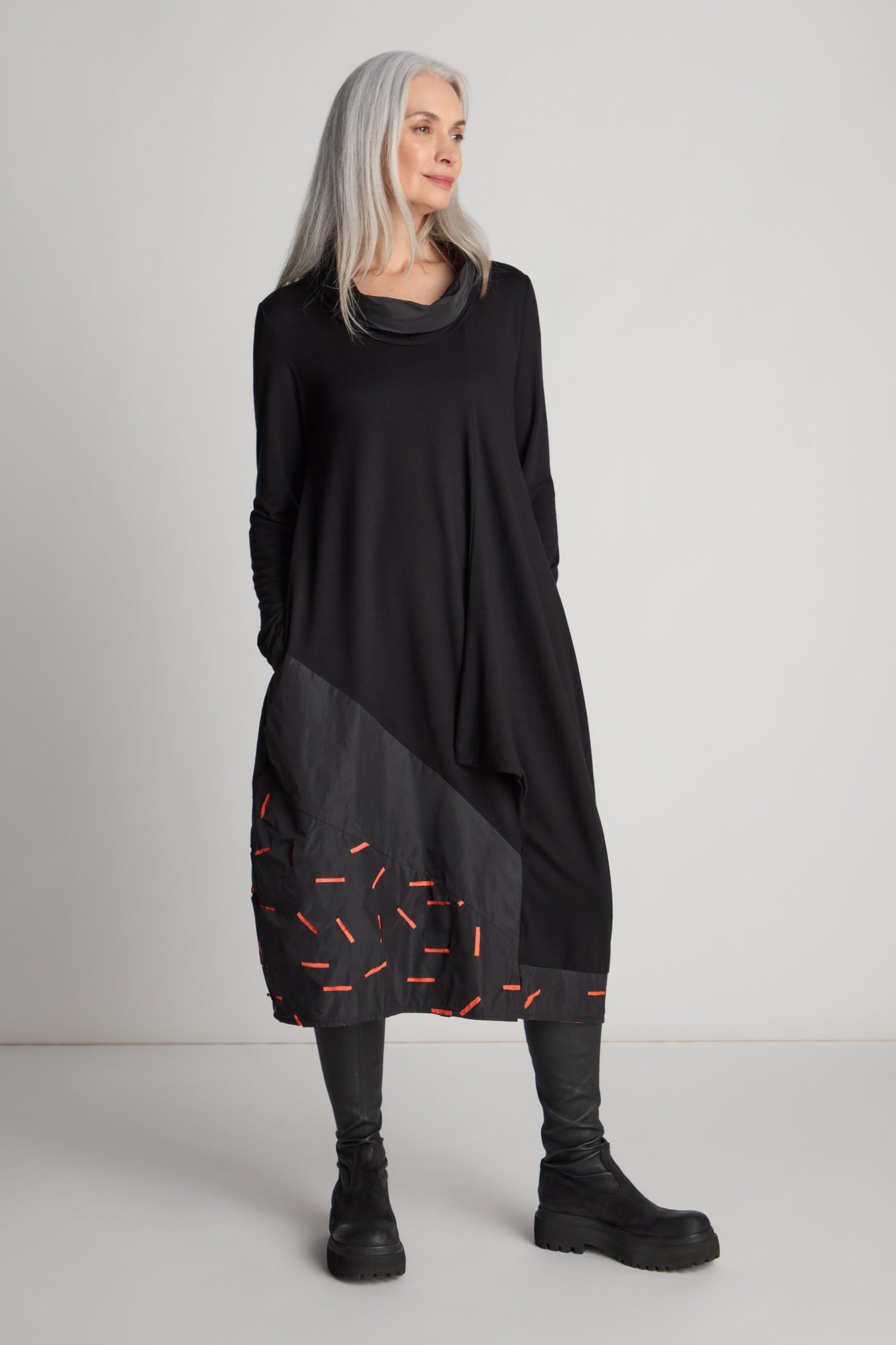 A woman with long gray hair stands against a neutral background, wearing a striking Cut Out Cowl Neck Dress as a statement piece, complemented by black leggings and black boots.