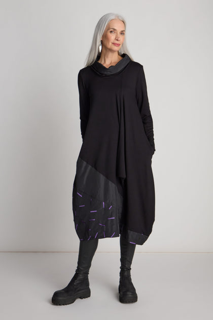 A woman with long gray hair stands against a white background, wearing a Cut Out Cowl Neck Dress by Luukaa—a true statement piece featuring a patterned lower half—paired with black leggings and black boots.