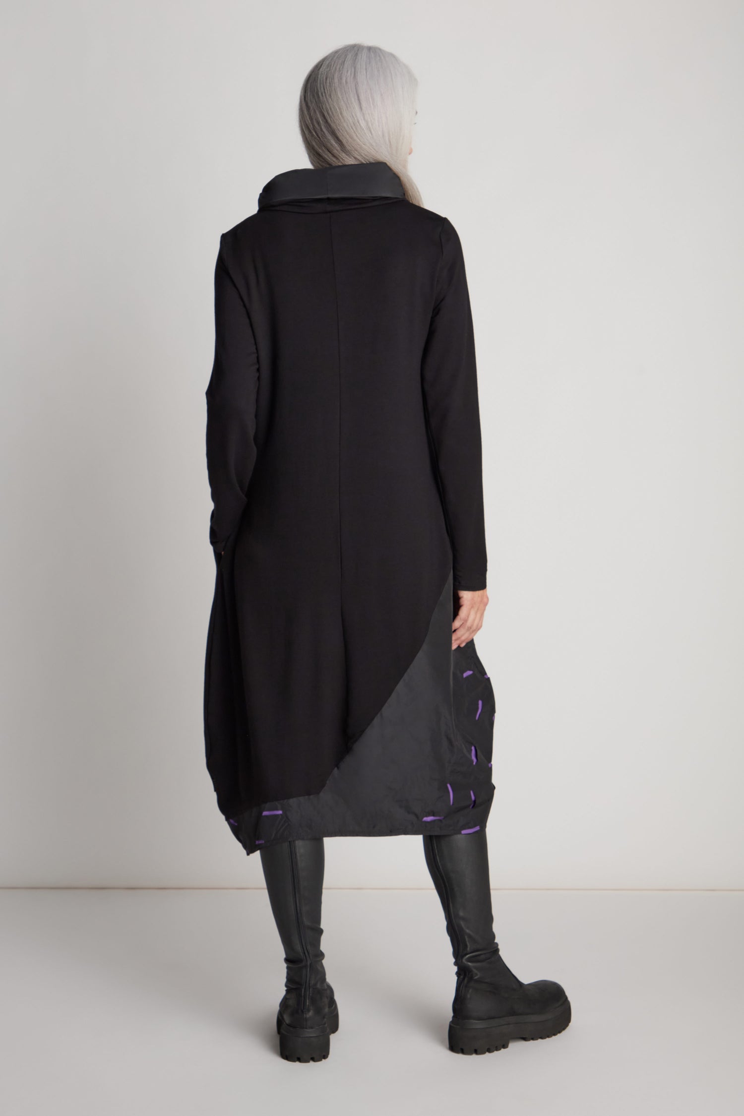 A person with long white hair stands facing away, wearing a Luukaa statement piece: the Cut Out Cowl Neck Dress paired with black leggings and boots.